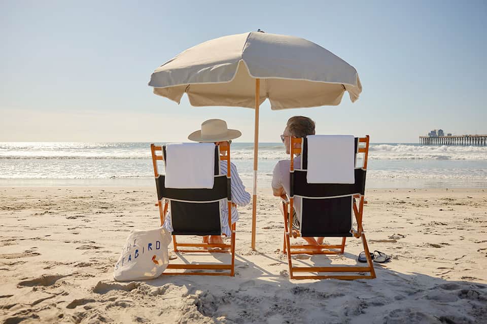 Couple relaxing under beach umbrella in chairs with ocean and pier in background
