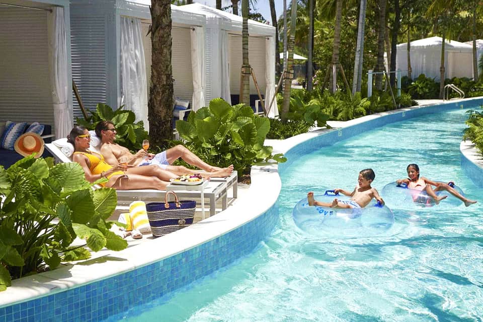 Family enjoying a curved resort pool surrounded by palm trees and cabanas with tropical landscaping