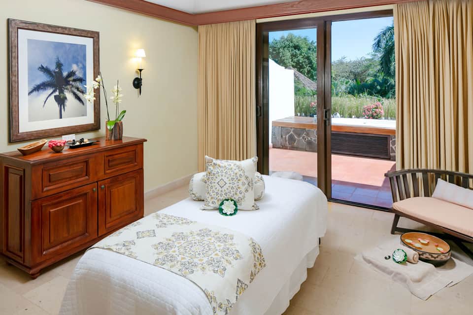 Spa treatment room with massage bed, palm artwork, wooden dresser, and glass doors opening to terrace garden