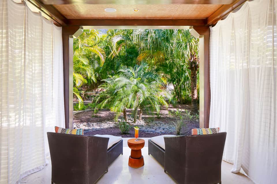 Covered outdoor seating area with wicker chairs overlooking lush tropical garden with palm trees