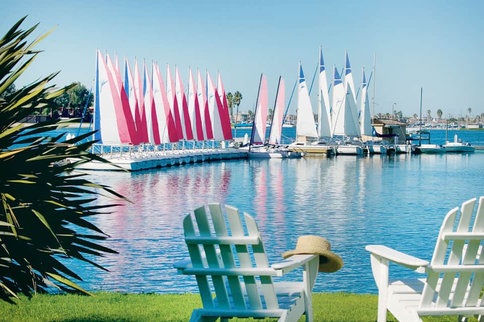 Sailboats with pink and white sails docked at marina, viewed from Adirondack chairs on waterfront lawn