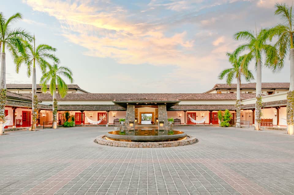 Traditional Asian-style courtyard with red doors, palm trees, and central fountain at dusk