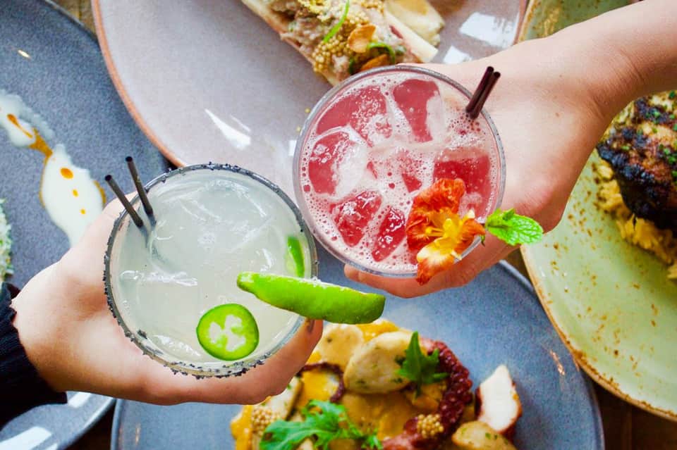 Two colorful cocktails with fresh garnishes held over plates of gourmet food at an outdoor dining table