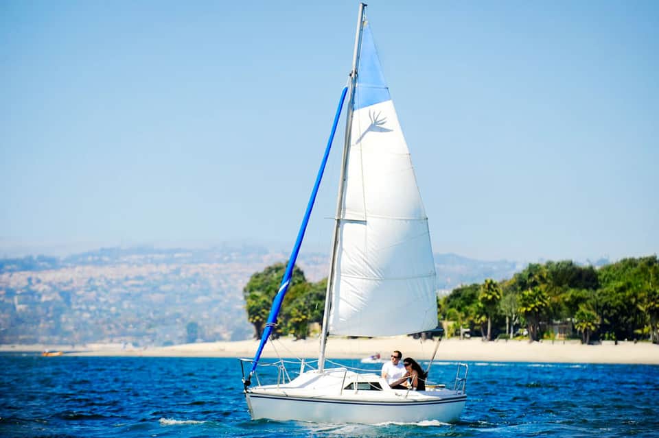 White sailboat with blue sail on ocean near sandy beach with palm trees and hills