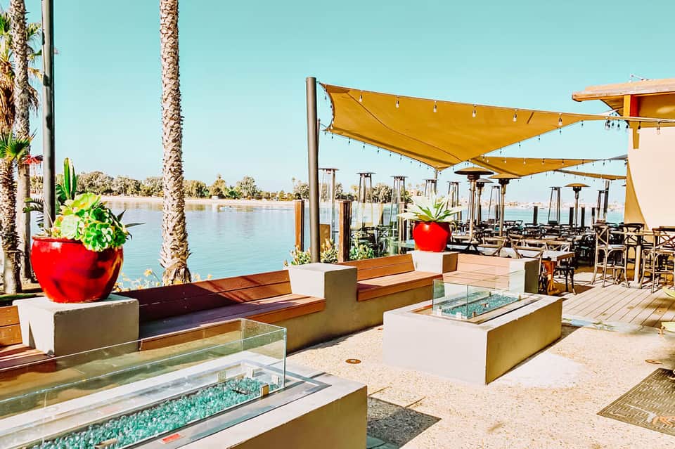 Waterfront dining patio with yellow umbrellas, fire pits, and palm trees overlooking a calm river