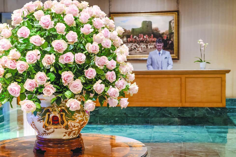 Luxurious pink rose arrangement in ornate vase at hotel reception desk with concierge in background
