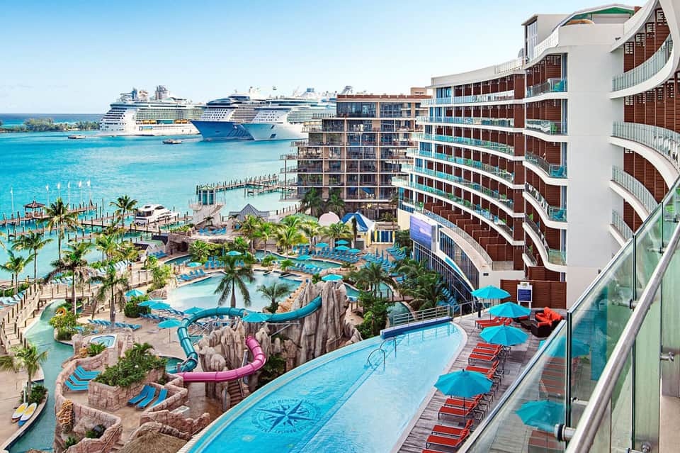 Beachfront resort with curved multi-story building, turquoise pools, water slides, and cruise ship in background