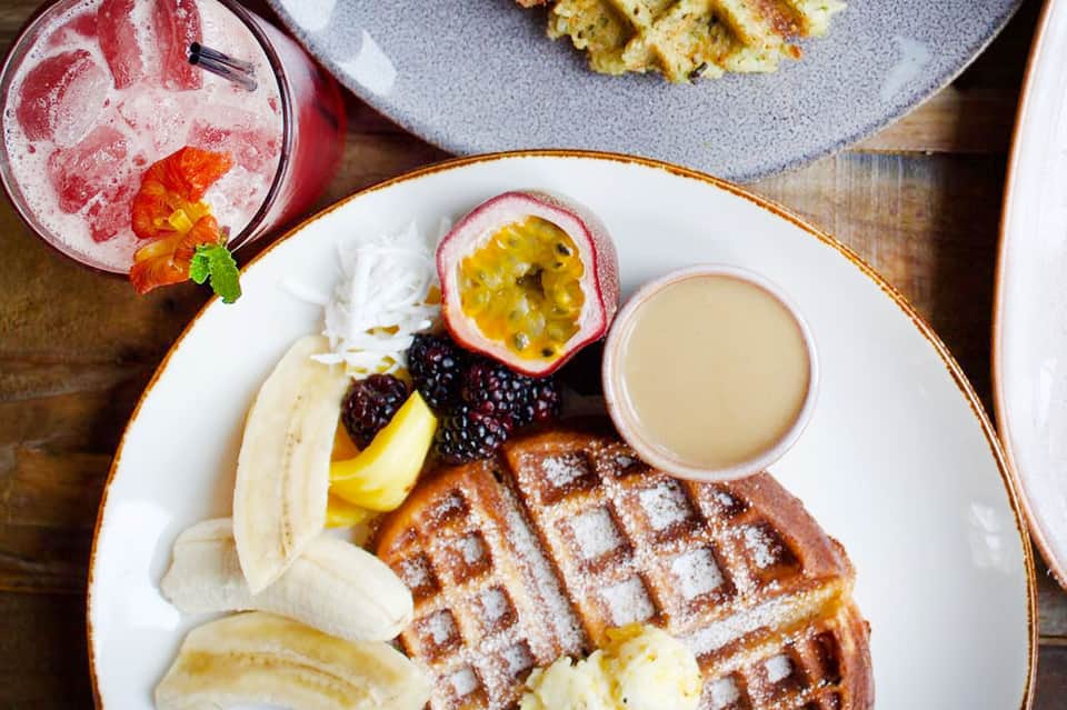 Waffle breakfast plate with fresh fruit, banana slices, berries, passion fruit, and cream sauce on white plate