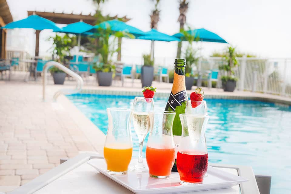 Poolside beverage station with champagne bottle and cocktails in glasses garnished with strawberries and tropical fruit.