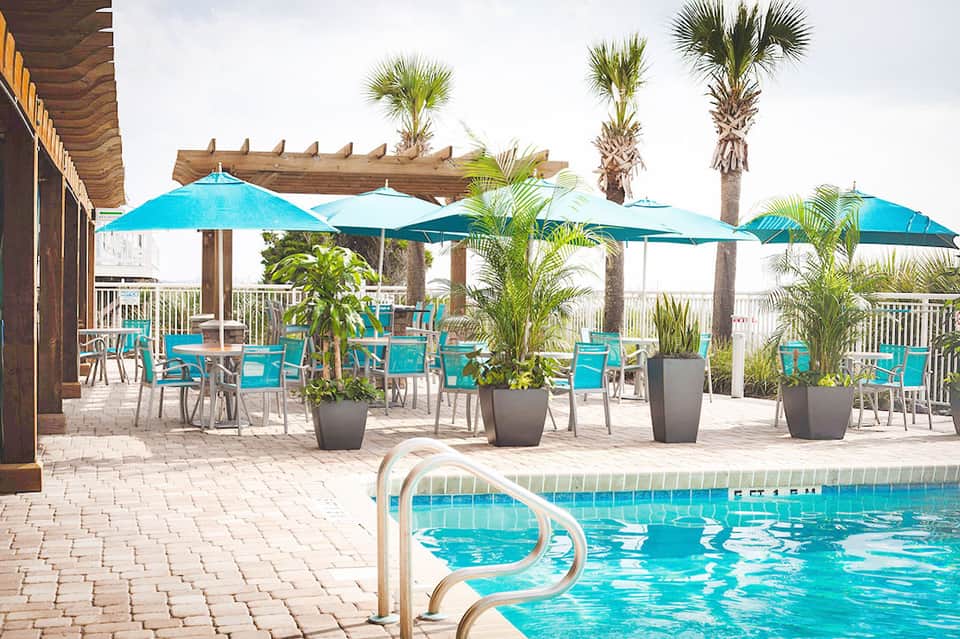 Poolside seating area with turquoise umbrellas, palm trees, and coastal views