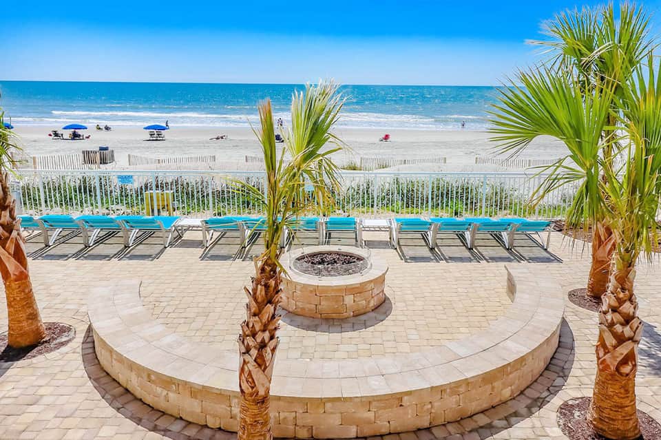 Beach boardwalk with blue loungers, palm trees, fire pit, and ocean view with white sandy beach and beachgoers