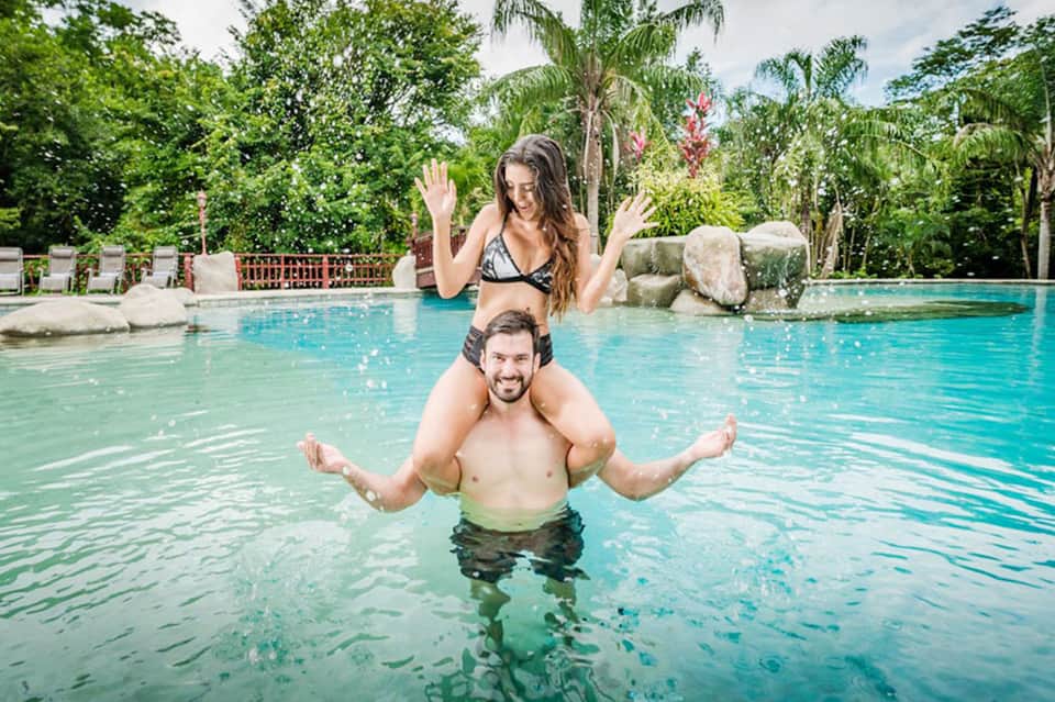 Couple playing in tropical resort pool surrounded by palm trees and lush greenery