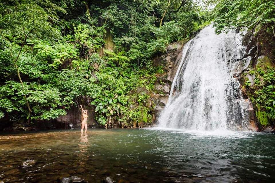 Person wading in tropical waterfall pool surrounded by lush green jungle vegetation
