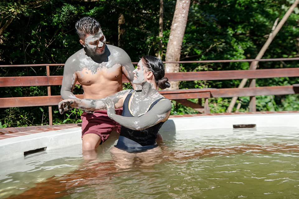 Couple applying mineral mud masks in natural hot spring pool surrounded by jungle