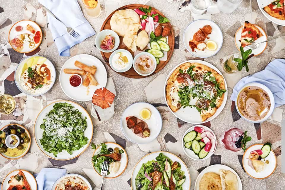 Overhead view of Mediterranean lunch spread with salads, pizza, hummus, and fresh vegetables on stone surface