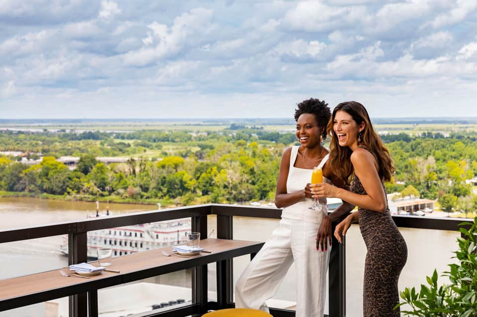 Two women on a riverside terrace enjoying drinks with expansive valley landscape views