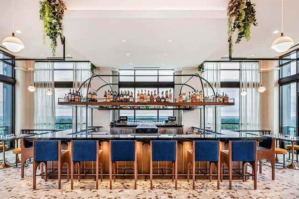 Modern beach bar with sand floor, wood bar, blue seating, hanging plants, and coastal views