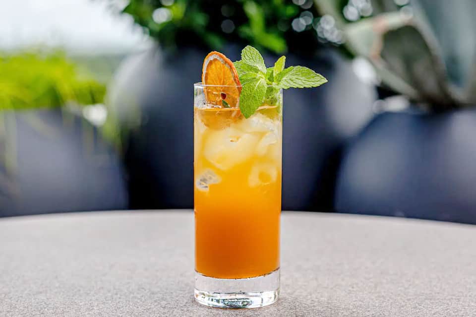 Orange cocktail with ice, mint garnish, and orange slice in tall glass by pool area