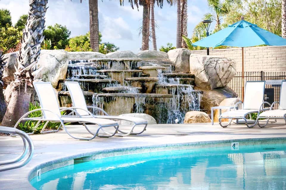 Resort pool with cascading waterfall, lounge chairs, palm trees, and blue umbrella on sunny day