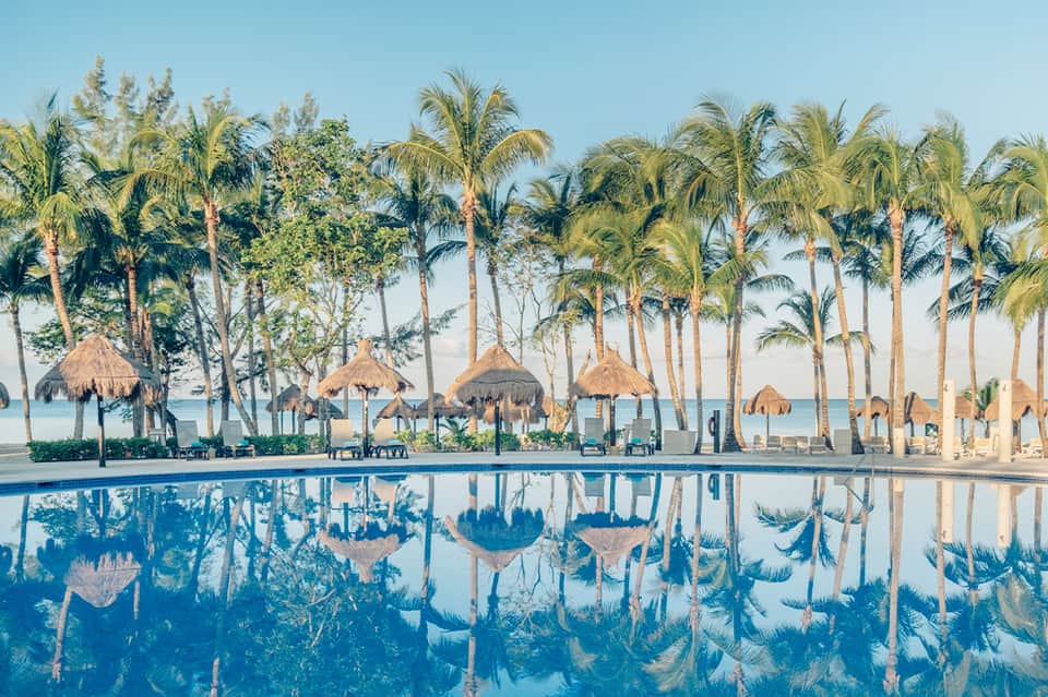 Resort pool with thatched umbrellas and palm trees reflecting in calm blue water at sunset
