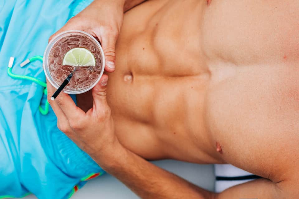 Shirtless man holding tropical cocktail with lime garnish by pool in bright sunlight