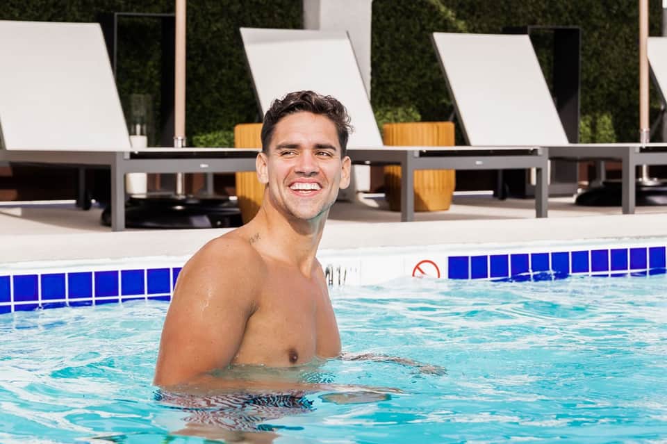 Man smiling in resort pool with lounge chairs and green hedges in background