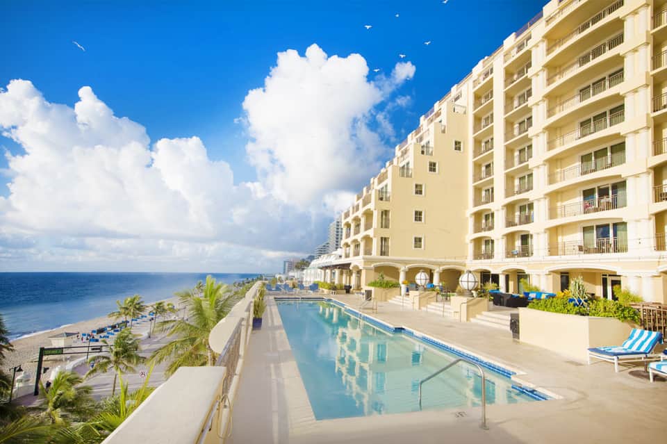 Beachfront resort with cream-colored building, oceanview pool, and palm trees under blue sky