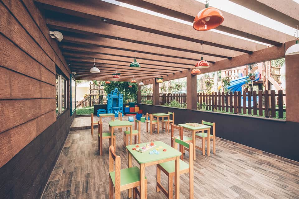 Indoor children's play area with colorful tables, chairs, and playground equipment under wooden beams