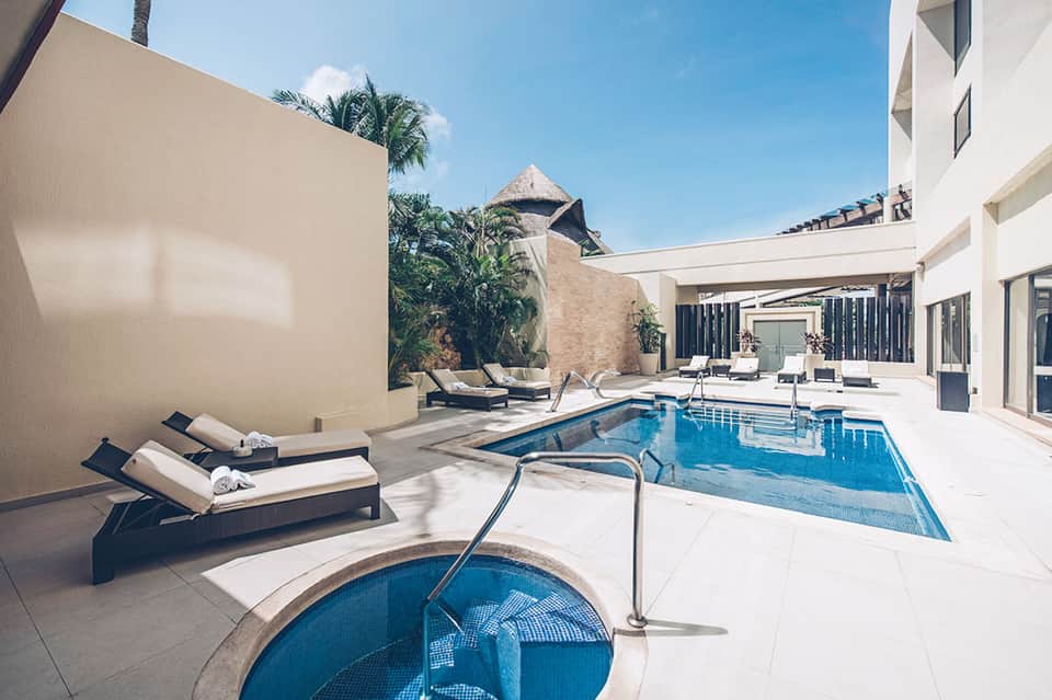 Modern white courtyard with rectangular pool, spa tub, lounge chairs, and thatched structures