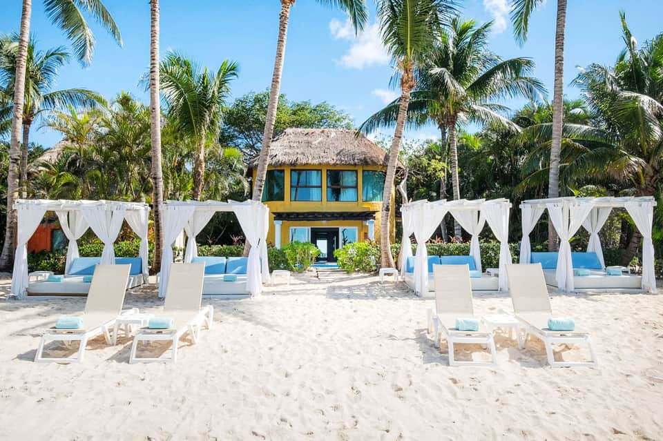 Tropical beach resort with white cabanas, lounge chairs, palm trees, and yellow building with thatched roof