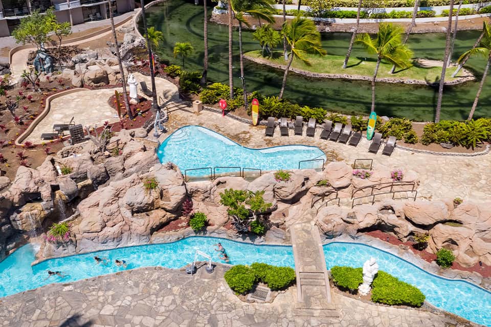 Resort pool area with turquoise water, rocky features, lounge chairs, and manicured green lawns overlooking lagoon