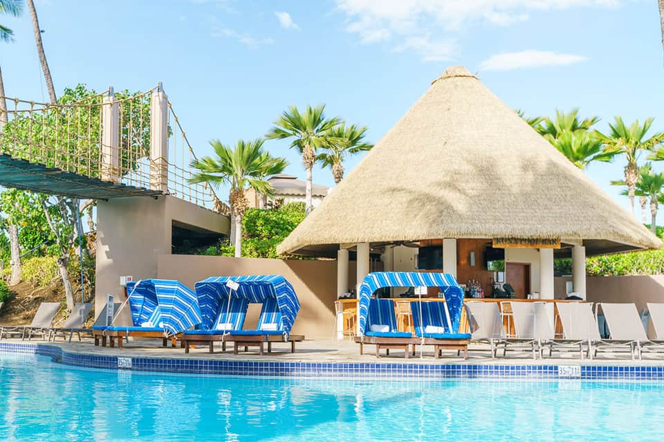 Resort pool area with thatched-roof cabana, blue loungers, and palm trees under clear sky