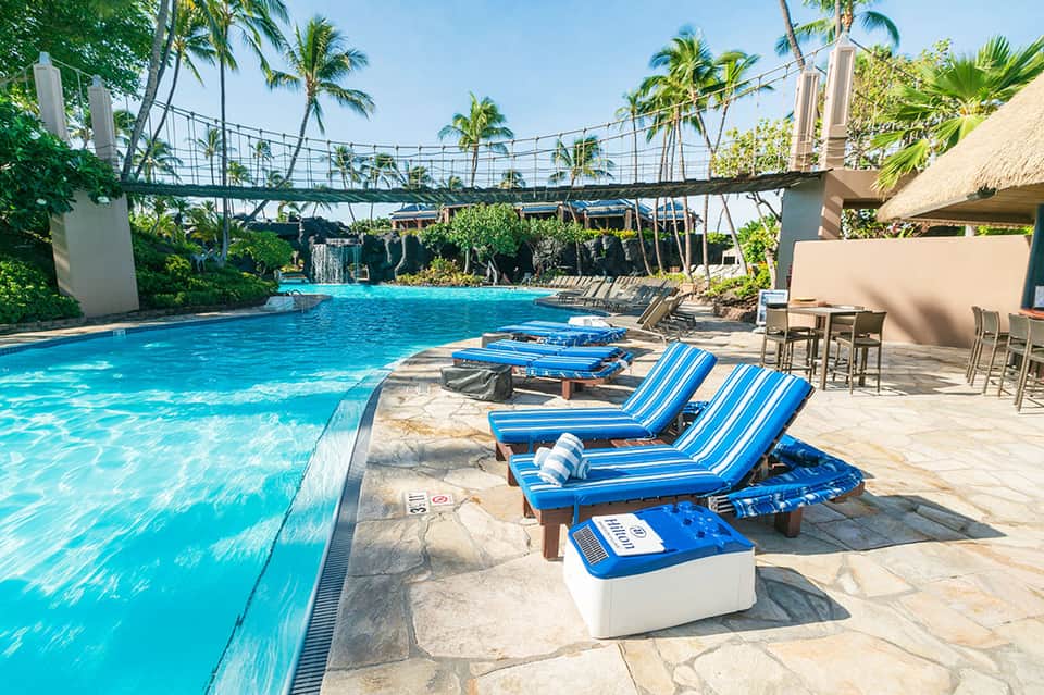 Tropical resort pool with blue striped loungers, palm trees, and suspension bridge