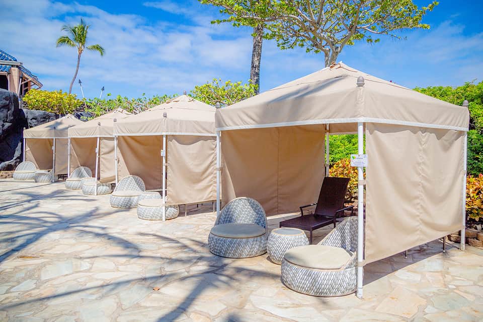 Row of beige canvas cabanas with lounge chairs on stone patio with palm trees and ocean views
