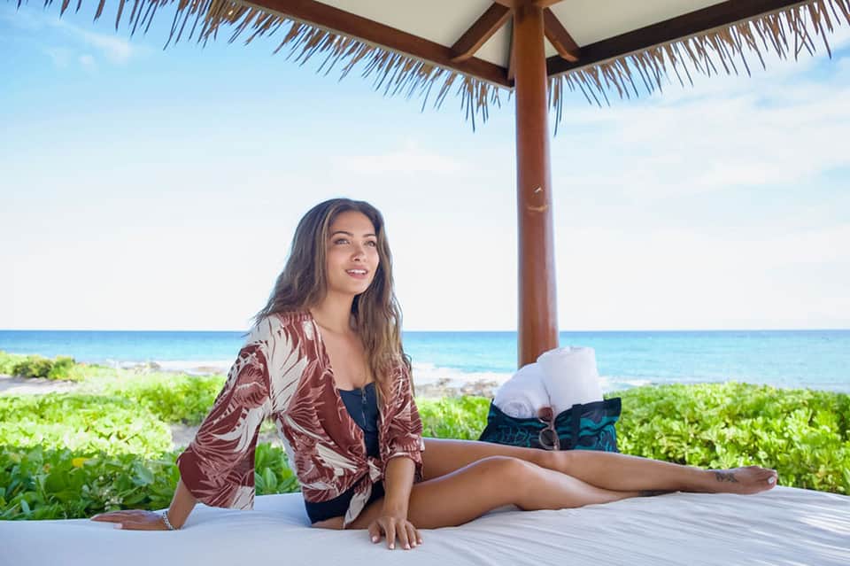 Woman in patterned jacket relaxing on beach platform under thatched umbrella with ocean view