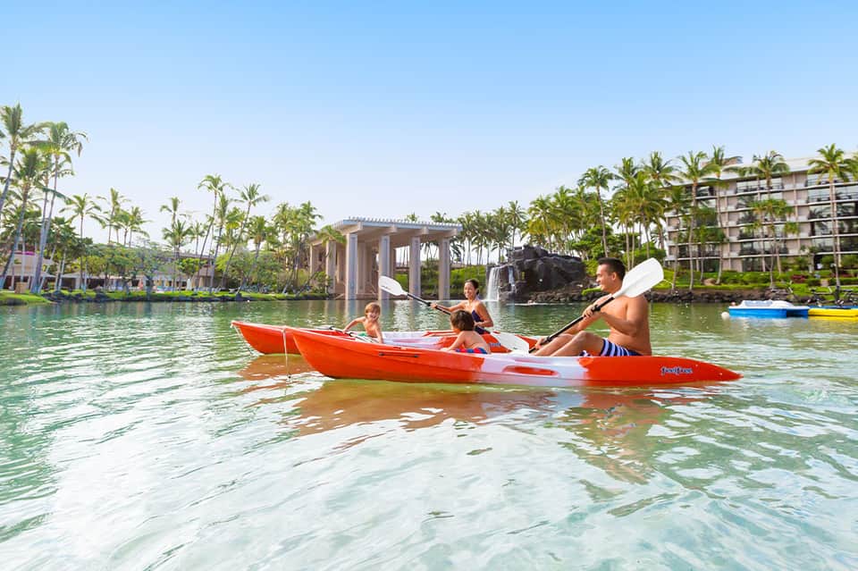 Family kayaking in shallow tropical waters near palm-lined resort with modern architecture
