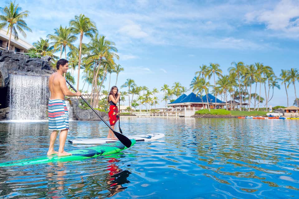 Man and woman paddleboarding on calm turquoise water with palm trees and beach buildings in background