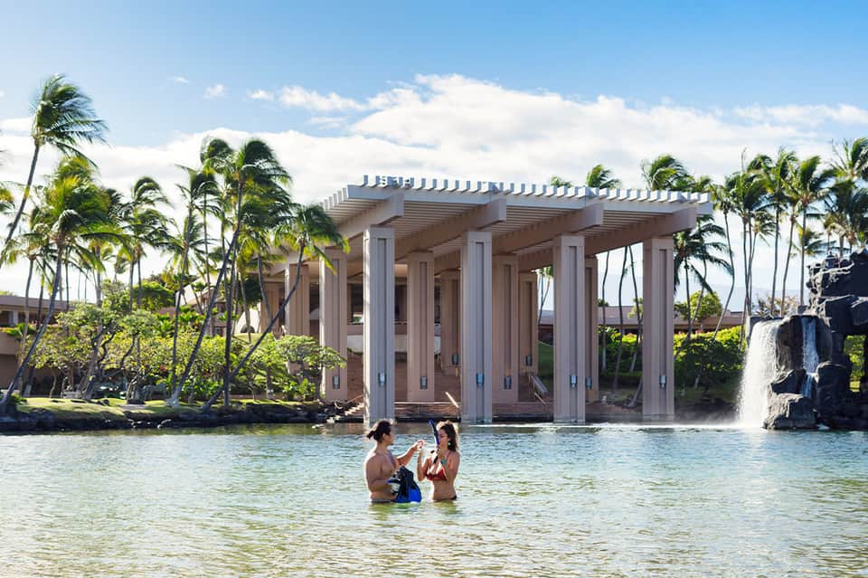 Family swimming in lagoon with large white columned structure and palm trees in background