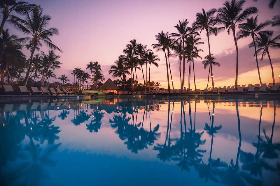 Tropical resort pool at sunset with palm trees reflected in still water at dusk
