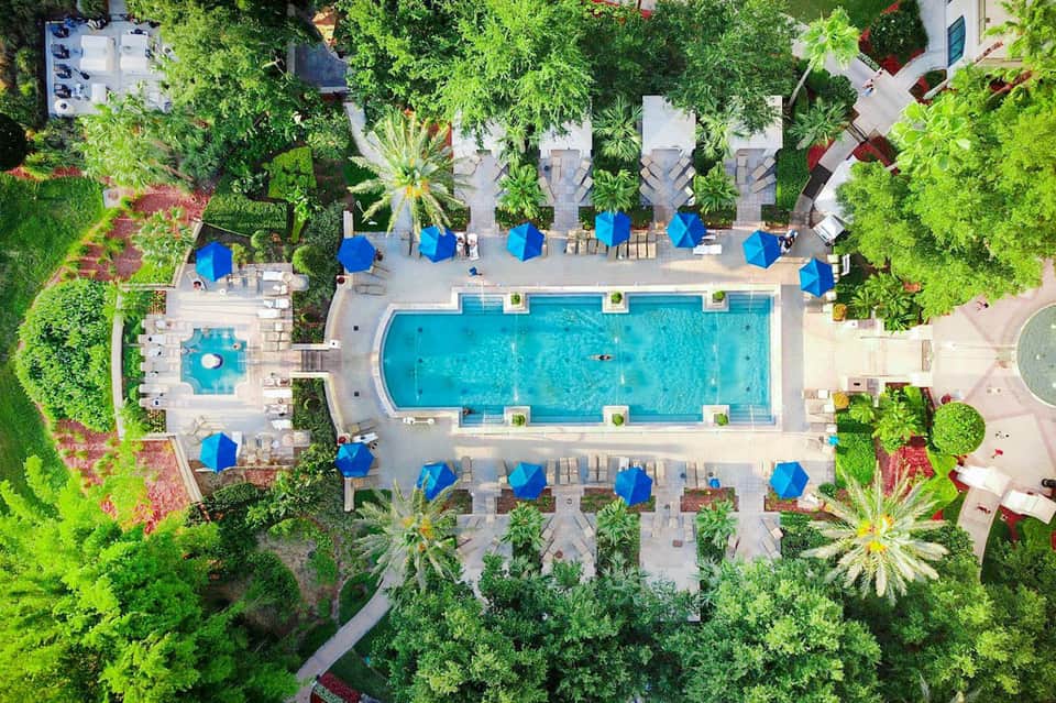 Aerial view of resort pool surrounded by blue umbrellas, loungers, and lush green landscaping