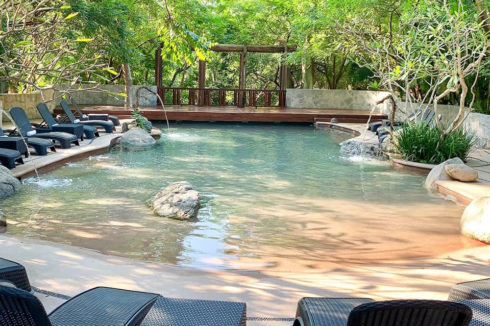Natural hot spring pool surrounded by lush trees with wooden deck and lounge chairs