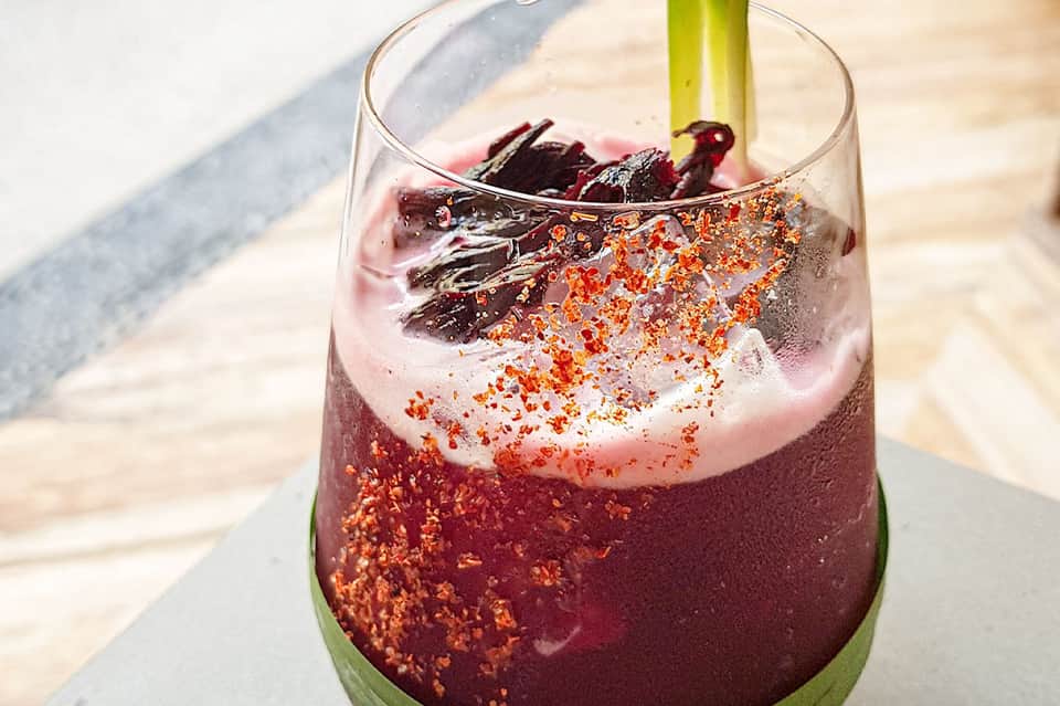 Tropical cocktail with pink foam, spicy rim, and herb garnish on white railing by beach