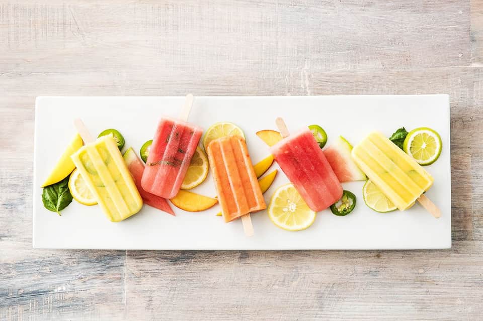 Colorful fruit popsicles and fresh fruit slices arranged on a white platter