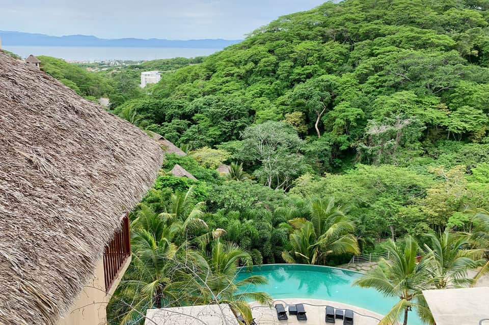 Aerial view of turquoise pool nestled in lush jungle valley with thatched-roof structures and ocean beyond