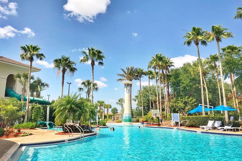 Tropical resort pool with turquoise water, palm trees, water slide, and white architecture under blue sky