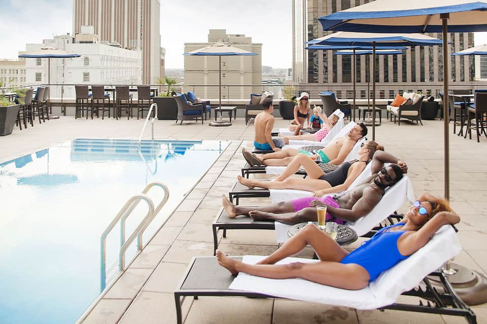Urban rooftop pool with multiple guests lounging on chairs under blue umbrellas