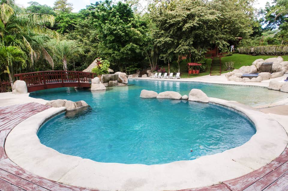 Resort swimming pool surrounded by lush greenery, white loungers, and wooden bridge