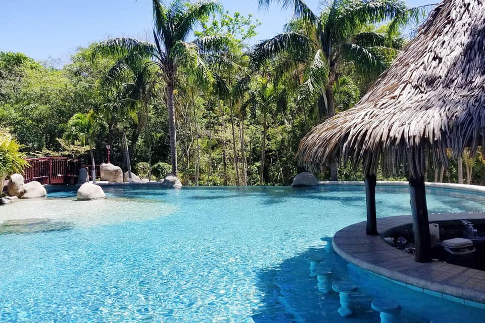 Tropical resort pool surrounded by lush palm trees and thatched-roof structures with clear blue water