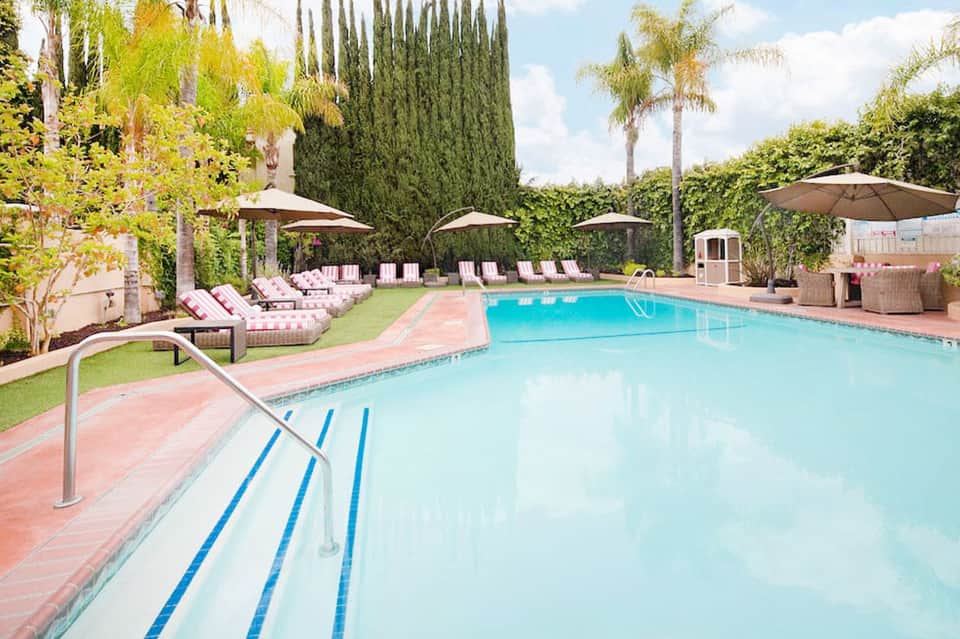 Resort swimming pool with lap lanes, pink and white lounge chairs under umbrellas, tropical landscaping with palm trees