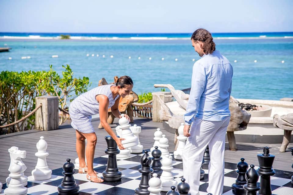 Couple playing oversized chess on oceanfront deck with turquoise water and horizon